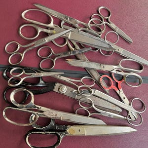 May include: A collection of various metal scissors in different sizes and styles, including some with black handles and one with red accents, arranged in a pile. The scissors feature sharp blades and finger loops, showing signs of wear.
