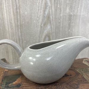 May include: A light gray ceramic gravy boat with a smooth, speckled finish. The vessel features a rounded body, a curved handle on one side, and a tapered pouring spout on the opposite side, creating a continuous, fluid silhouette for serving.