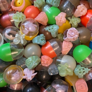 May include: A collection of colorful plastic capsules filled with various small toys, including skulls, rings, and other whimsical designs. The capsules are a mix of clear, opaque, and translucent colors.