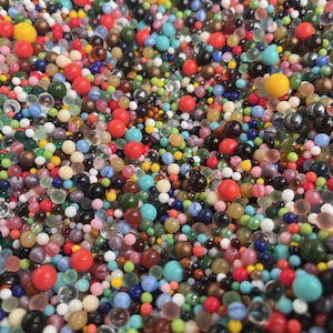 May include: A dense collection of small, spherical beads in a wide variety of colors including red, yellow, blue, green, black, white, and clear. The beads vary in size and finish, ranging from opaque and matte to translucent and glossy textures.