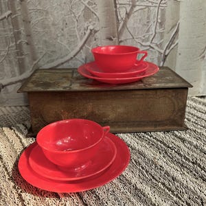 May include: Two sets of bright red teacups and matching saucers. Each set consists of a rounded cup with a small handle and a circular saucer. The vibrant red color is consistent across all pieces, which feature a smooth, solid finish.