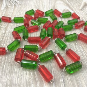 May include: A necklace composed of alternating red and green cylindrical beads. The beads are strung together with silver-colored metal links. The beads are translucent, and the necklace is lying on a light-colored wooden surface.