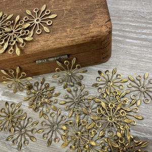 May include: A collection of antique brass snowflake charms scattered on a wooden surface and a wooden box. The charms have a detailed, openwork design with delicate petal-like shapes and small circular accents, perfect for jewelry making.