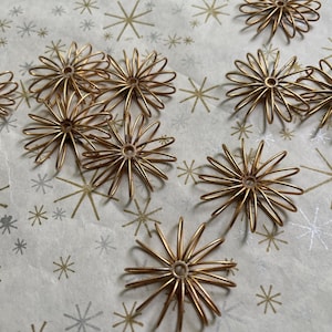 May include: A collection of gold-colored wire starburst ornaments. The ornaments are arranged on a white and gold patterned surface. The starbursts have a central ring with radiating wire spokes, creating a delicate, festive appearance.