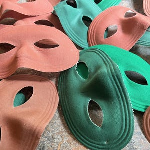 May include: A collection of fabric eye masks in shades of coral and green. The masks have oval eye holes and are layered with a textured, ridged design. These masks are suitable for costume parties or theatrical events.