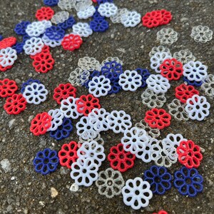 May include: A collection of red, white, and blue plastic flower-shaped embellishments. The embellishments are scattered on a gray surface.
