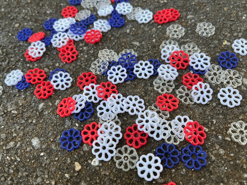 May include: A collection of red, white, and blue plastic flower-shaped embellishments. The embellishments are scattered on a gray surface.