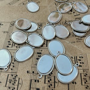 May include: A collection of silver oval-shaped metal blanks with a raised edge. The blanks have a hole at the top for hanging.
