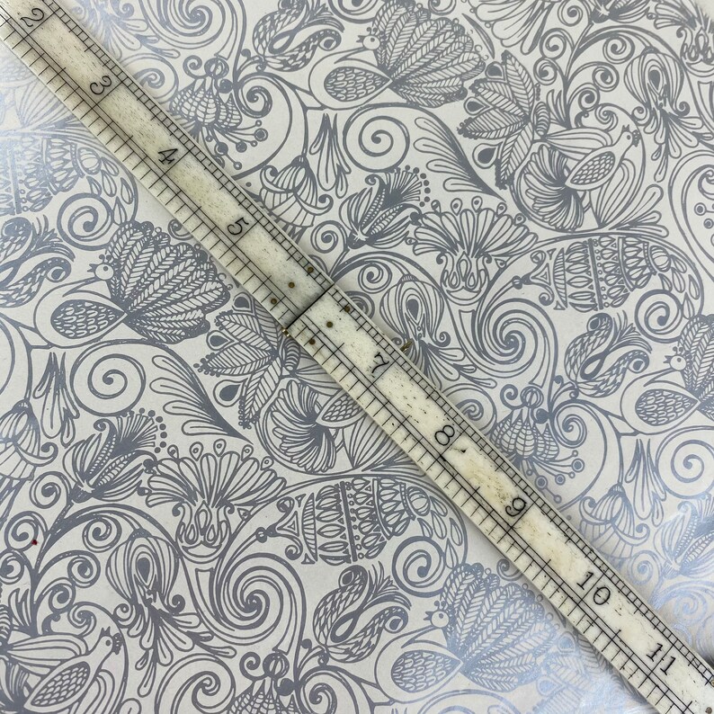 May include: A close-up of a white background with a silver-grey floral pattern. A ruler is placed diagonally across the image, with the numbers 3 through 11 visible.