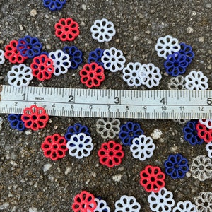 May include: A collection of red, white, and blue plastic flower-shaped embellishments. The embellishments are scattered on a gray surface with a ruler measuring 5 inches.