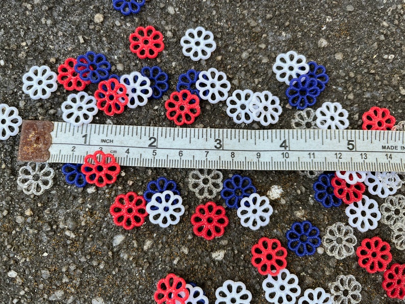 May include: A collection of red, white, and blue plastic flower-shaped embellishments. The embellishments are scattered on a gray surface with a ruler measuring 5 inches.