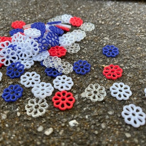 May include: A pile of small, decorative, plastic flowers in red, white, and blue. The flowers are scattered on a gray, textured surface.