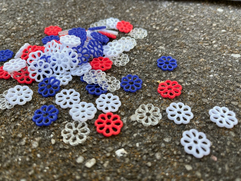 May include: A pile of small, decorative, plastic flowers in red, white, and blue. The flowers are scattered on a gray, textured surface.