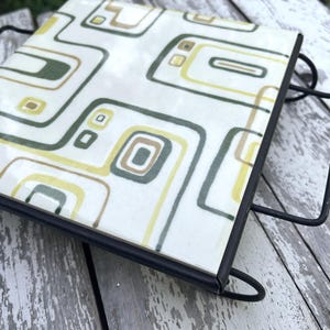 May include: A square ceramic tile trivet featuring a repeating pattern of olive green, mustard yellow, and brown rounded rectangular shapes on a cream background, set within a black metal wire frame with integrated side handles.