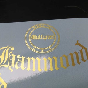 May include: Close-up of a light blue surface with gold foil lettering. The text includes "Made in U.S.A." and the name "Hammond" in an ornate font. A circular logo reads "Multiplex".