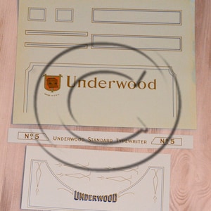 May include: A vintage Underwood typewriter label with a gold and blue design. The label features the text "Underwood Standard Typewriter" and the number "No. 5".