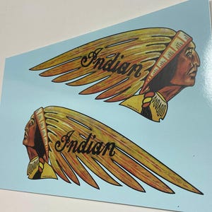 May include: Two vintage Indian Motorcycle decals featuring a Native American head with a feathered headdress and the word "Indian" in a stylised font. The decals are on a light blue background.
