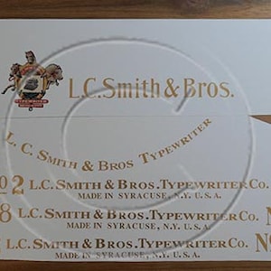 Puede incluir: Una etiqueta vintage de la empresa L.C. Smith & Bros. Typewriter Company con letras doradas sobre fondo blanco. La etiqueta dice "NO 2 L.C. Smith & Bros. Typewriter Co. NO 2 Made in Syracuse, N.Y. U.S.A."