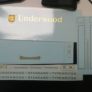 May include: A collection of light blue and gold Underwood typewriter decals. The decals include the Underwood logo, measurements in inches, and text that reads "Underwood Standard Typewriter".