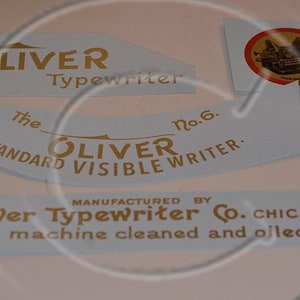 Könnte beinhalten: Vier vintage Etiketten aus blauem und weißem Papier mit goldenen Schriftzügen für eine Oliver Schreibmaschine. Die Etiketten lauten "The Oliver Typewriter", "The Oliver Standard Visible Writer No. 6" und "The Oliver Typewriter Co. Chicago, U.S.A. Keep machine cleaned and oiled."