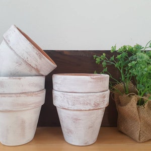 May include: Set of four whitewashed terracotta flower pots, stacked and arranged on a wooden surface. The pots have a rustic, distressed look and are perfect for adding a touch of farmhouse charm to any home.