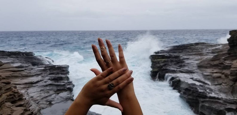 Pu&ograve; includere: Due mani con anelli d'oro sullo sfondo di una costa rocciosa con onde che si infrangono.