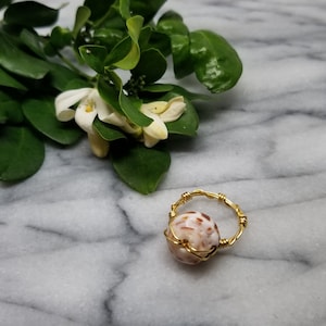 May include: A handmade ring with a gold-colored wire band. The ring features a round, light brown and white stone, wrapped in the same gold wire. Green leaves and white flowers are in the background, set against a marble surface.