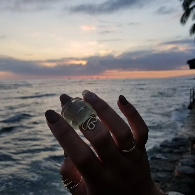 Pu&ograve; includere: Una mano con un anello con una pietra di vetro trasparente e una fascia dorata. La mano &egrave; sullo sfondo di un tramonto sull'oceano.