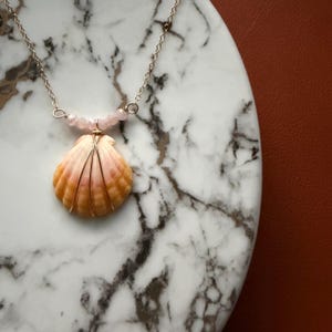May include: A seashell pendant necklace with a silver chain and pink gemstone beads. The seashell is a light orange color with gold wire wrapping. The necklace is displayed on a marble surface.