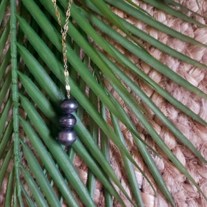May include: A gold chain necklace with three black pearls hanging from it. The necklace is on a green leafy background.