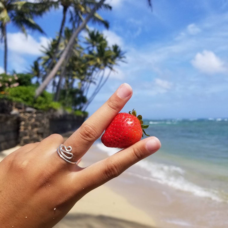 Pu&ograve; includere: Una mano che tiene una fragola rossa davanti a uno scenario di spiaggia con palme e cielo blu.