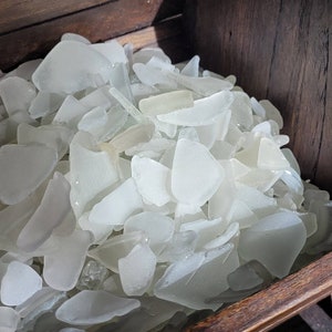 May include: A wooden box filled with a large collection of white sea glass. The glass is smooth and translucent, with a variety of shapes and sizes.
