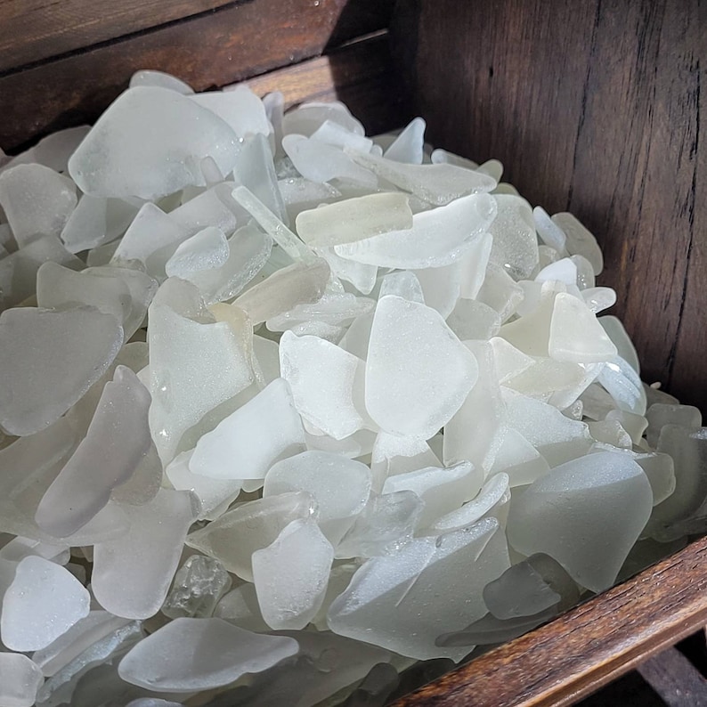 May include: A wooden box filled with a large collection of white sea glass. The glass is smooth and translucent, with a variety of shapes and sizes.