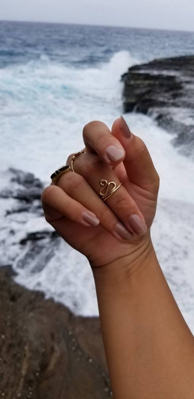Pu&ograve; includere: Primo piano di una mano che indossa due anelli d'oro. L'anello sull'indice &egrave; una semplice fascia, mentre l'anello sul medio &egrave; un design a doppio anello. La mano &egrave; sullo sfondo di una spiaggia con onde che si infrangono.