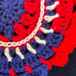 May include: Close-up of a crocheted round doily with a red, white, and blue color scheme. The doily has a scalloped edge and a center with a starburst pattern.