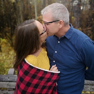 May include: A couple kissing outdoors. The woman wears a yellow top and is wrapped in a red and black plaid blanket. The man wears a blue button-down shirt. The background is a blurred forest scene.