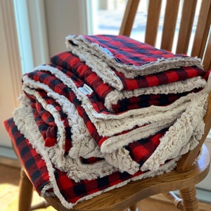 May include: A stack of red and black plaid blankets with a white sherpa lining. The blankets are folded and stacked on a wooden chair.