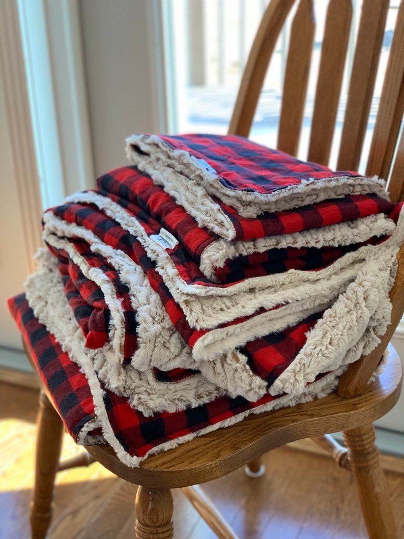 May include: A stack of red and black plaid blankets with a white sherpa lining. The blankets are folded and stacked on a wooden chair.