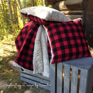 May include: Two red and black plaid throw pillows with a white faux fur pillow on top. The pillows are sitting on a gray wooden crate.