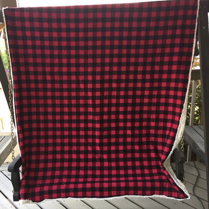 May include: A red and black buffalo plaid blanket with a white, fluffy sherpa lining. The blanket is draped over a black swing, showcasing the contrasting textures and colors. The plaid pattern is a classic design.