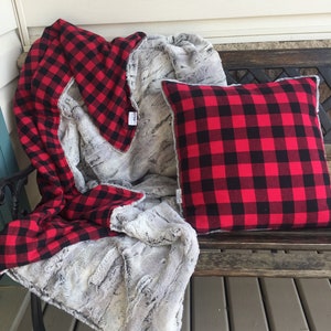 May include: A red and black buffalo plaid throw blanket with a faux fur backing. The blanket is folded and draped over a wooden bench with a matching pillow.