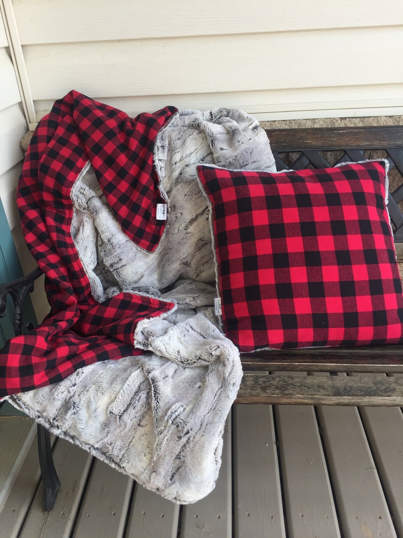 May include: A red and black buffalo plaid throw blanket with a faux fur backing. The blanket is folded and draped over a wooden bench with a matching pillow.