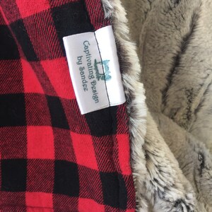 May include: A red and black plaid fabric with a white tag that reads "Captivating Design by Sandee". The tag is attached to a gray and white faux fur fabric.