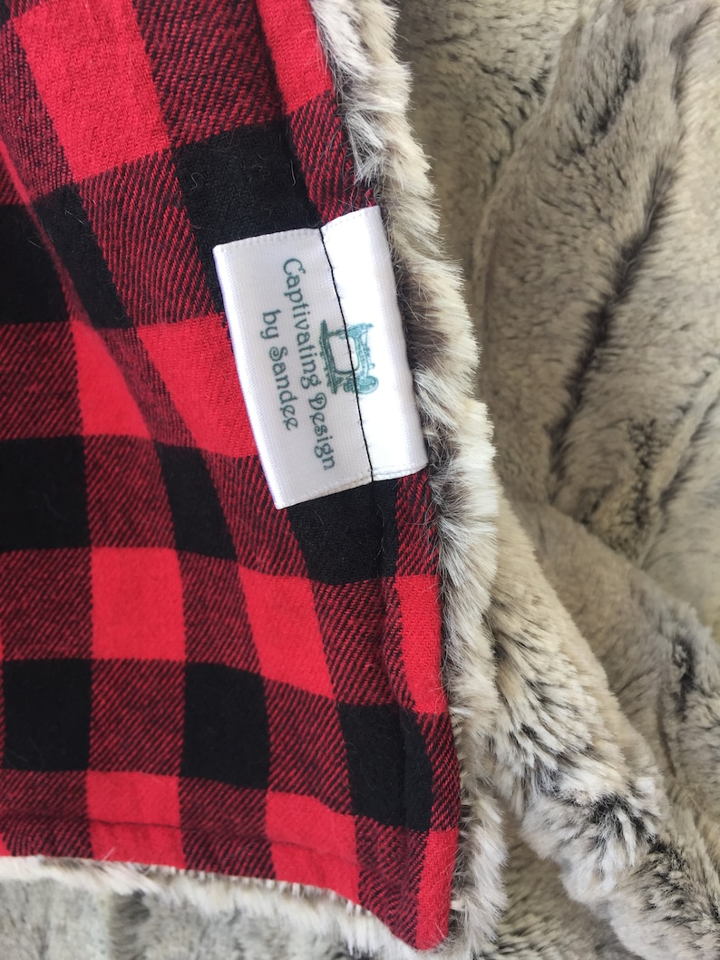 May include: A red and black plaid fabric with a white tag that reads "Captivating Design by Sandee". The tag is attached to a gray and white faux fur fabric.