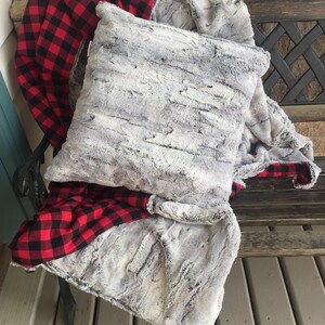 May include: A gray faux fur throw blanket with a red and black plaid flannel lining. The blanket is folded and draped over a wooden bench.