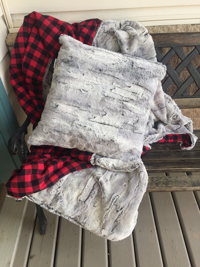 May include: A gray faux fur throw blanket with a red and black plaid flannel lining. The blanket is folded and draped over a wooden bench.