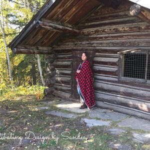 May include: A woman stands in front of a rustic wooden cabin, wearing a red and black plaid blanket. The cabin has a wooden door and a window with a wire mesh screen. The woman is standing on a stone path in front of the cabin.