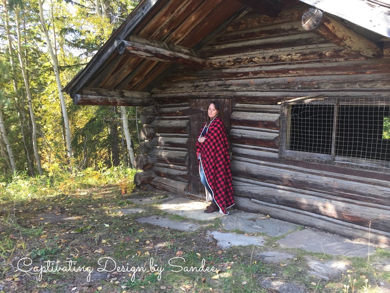 May include: A woman stands in front of a rustic wooden cabin, wearing a red and black plaid blanket. The cabin has a wooden door and a window with a wire mesh screen. The woman is standing on a stone path in front of the cabin.