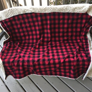 May include: A red and black buffalo plaid blanket with a cream-colored, plush sherpa lining. The blanket is draped over a wooden bench, showcasing the contrasting textures and colors. The plaid pattern is a classic design.