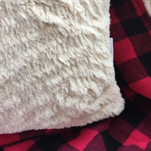 May include: A cream-colored faux fur throw pillow with a soft, fluffy texture. The pillow is lying on a red and black plaid fabric.